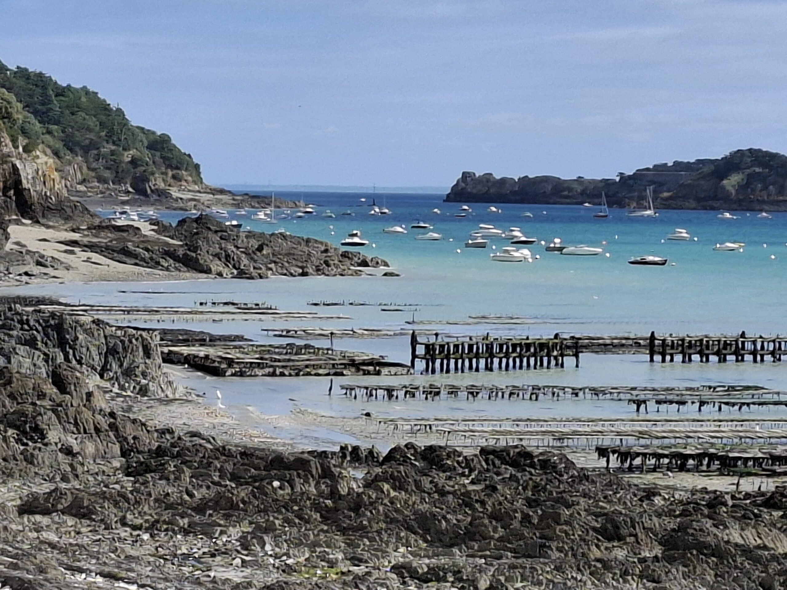 Vue de la côte depuis le GR34 sur la commune de Cancale. L'eau est turquoise, quelques bateaux sont au mouillage et on aperçoit le début des parcs à huîtres, légèrement recouverts par la marée.